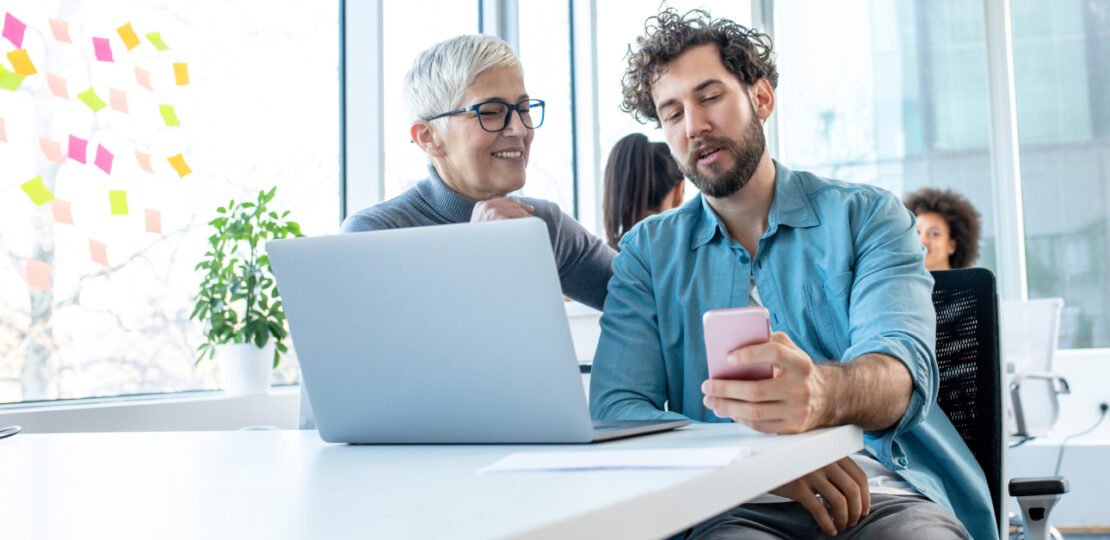 A relaxed office atmosphere, work break. A man is holding a phone and talking to a senior colleague. There are two more women behind and there are sticky notes on a large window.